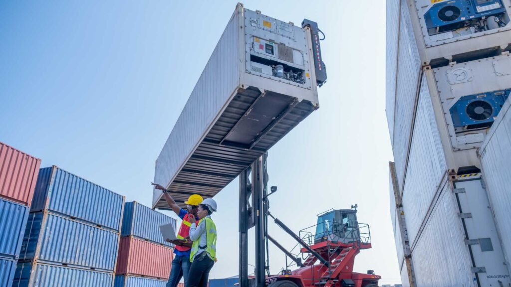 Muletto in funzione solleva container mentre un caposquadra e un tecnico supervisionano le operazioni di carico in area logistica